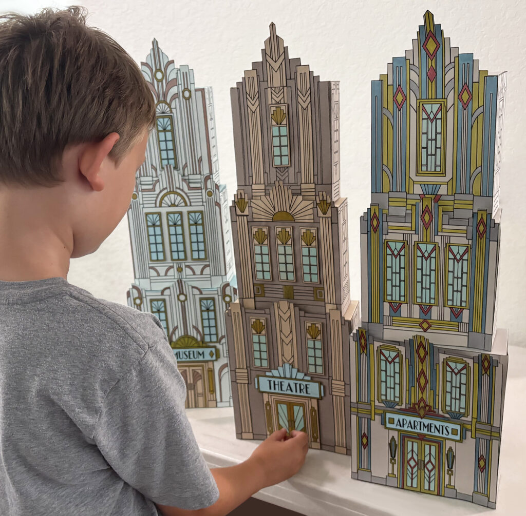 Child standing beside three completed Art Deco paper skyscrapers, showing the large scale of the approximately 26-inch-tall buildings.