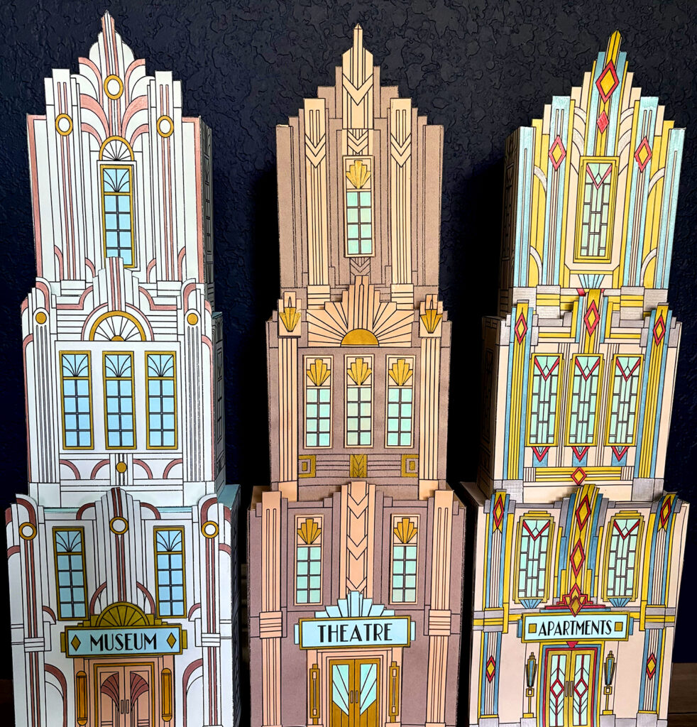 Three completed Art Deco paper skyscrapers with stepped rooflines, geometric windows, and colorful architectural details.